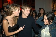 two girls and a boy laughing together in a crowded room with other people looking on