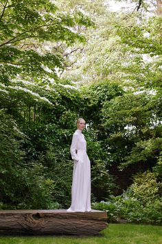 a woman in white dress standing on top of a wooden log with trees behind her