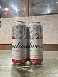 two cans of budweiser beer sitting on top of a marble table in front of a window