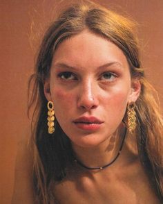 a woman with long hair and earrings on her neck looking at the camera while wearing gold jewelry