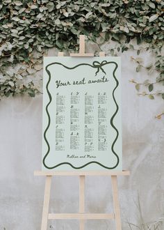 an easel with a seating chart on it in front of a wall covered in greenery