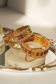 Introducing the Amika Bangle Set (Set of 2), a stunning addition to any jewelry collection. These gold-plated bangles feature intricate designs adorned with pearls and vibrant multicolor stones, including green and ruby accents. The detailed floral and petal patterns exude elegance and sophistication, making these bangles & bracelets set perfect for desi weddings and special occasions. The Amika Bangle Set showcases the timeless beauty of Navratan craftsmanship. The bangles are designed with an