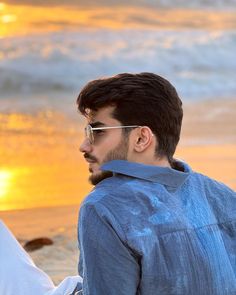 a man wearing sunglasses standing on the beach at sunset with his hand in his pocket