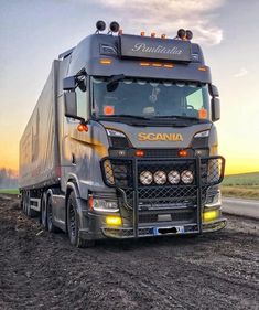 a large truck driving down a dirt road next to a green field at sunset with the sun setting behind it