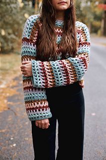 a woman standing on the side of a road wearing a crocheted sweater and black pants