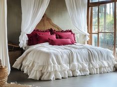 a bed with white ruffled bedspread and red pillows in front of a window