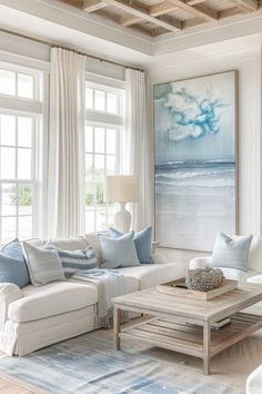 a living room filled with white furniture and blue pillows on top of a rug in front of two large windows