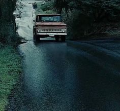 an old pickup truck driving down a wet road in the dark with trees on either side