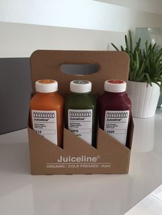 three bottles of juice sitting in a box on a counter top next to a potted plant