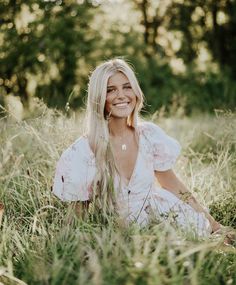 a woman sitting in tall grass smiling for the camera with trees in the back ground behind her