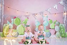 two babies sitting in front of cactus themed backdrop with cake and cupcakes for their first birthday