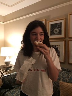 a woman is eating a doughnut while standing in front of a couch with pictures on the wall