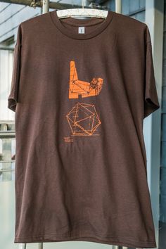 A brutalist T-shirt in size M. The color is brown, with a design of the church and a climbing frame in the Dautenbach in Siegen printed in orange. Made with love for architecture. Unique piece. 100% cotton SCREEN PRINTING, ART Brown Cotton Top With Graphic Design, Brown Cotton Graphic Design T-shirt, Architecture Unique, Screen Printing Art, Climbing Frame, The Church, Climbing, Unique Pieces, Screen Printing