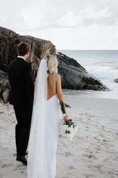 a bride and groom standing on the beach looking out at the ocean while holding hands