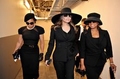 three women wearing hats and black outfits walking down a hall way with other people in the background