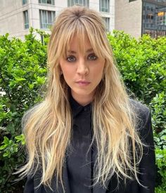 a woman with long blonde hair standing in front of bushes