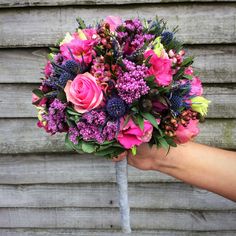 a bridal bouquet with pink roses and purple thistles is held by a hand