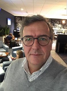 a man with glasses is taking a selfie in an office setting while people are sitting at tables and eating