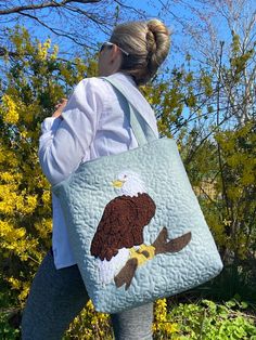 "This handmade bag has a beautiful bold Bald Eagle perched on a branch. This bag was made in mind for all of those eagle nest followers. So many famous nest cams are watched today. I have followed several nest over the years and have been inspired to make these bags. This bag makes a great gift and will last for many years.  The bag is approximately 13\"x 14\". It is extremely durable and lined. It has 3 inside pockets. It also has a magnetic clasp that opens and closes easily. The bag has nice long padded handles approximately 27 \" long. The front of the bag is quilted. Each bag I make is one of a kind because it is done freehand by me. The bags are also hand pieced and stitched in our smoke free home. Free shipping included with this purchase." Quilted Rectangular Bag For Gifts, Quilted Rectangular Bag For Gift, Quilted Pouch Bag As Gift, Quilted Rectangular Bag As Gift, Quilted Pouch Bag For Gift, Rectangular Quilted Bag For Gifts, Rectangular Quilted Bag As Gift, Eagle Quilt, Eagle Nest