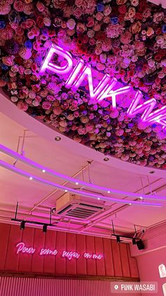 the pink neon sign is hanging from the ceiling in front of a wall of flowers