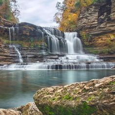 a large waterfall in the middle of a river surrounded by rocks and trees with fall foliage on either side