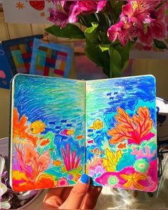 someone is holding up an open notebook with flowers in the background and watercolors on it