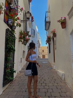 a woman is standing in an alleyway with flowers on the windows and potted plants