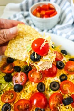 a person holding a tortilla chip with tomatoes and olives on it in front of a bowl of salad