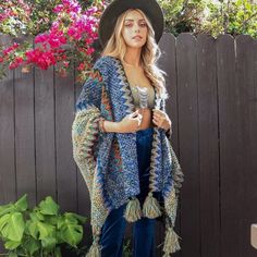 a woman standing in front of a fence wearing blue pants and a crocheted shawl