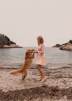 a woman is playing with her dog on the beach in front of some water and trees