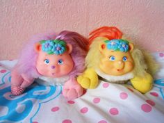 two troll dolls sitting on top of a bed next to each other, one with pink and blue hair