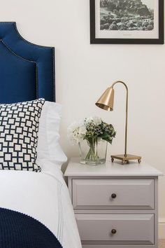 a nightstand with flowers on it next to a blue headboard and white bedding