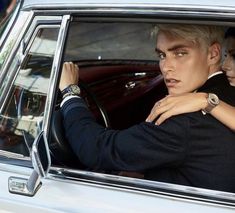 two people sitting in a car, one is holding the steering wheel and the other is wearing a suit