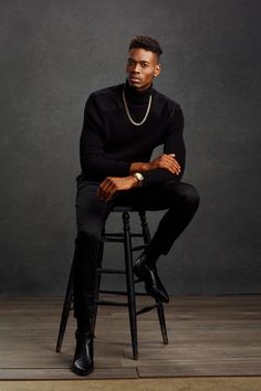 a man sitting on top of a wooden stool wearing a black sweater and pearls necklace