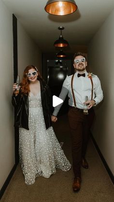 a man and woman walking down a hallway holding wine glasses in their hands while wearing eyeglasses