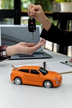 two people exchanging keys to an orange toy car on a table next to a laptop