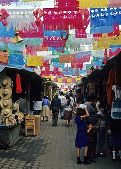 many people are walking through an outdoor market with lots of colorful items hanging from the ceiling