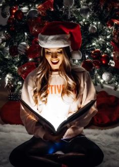 a woman is sitting in front of a christmas tree and reading a book while wearing a santa hat