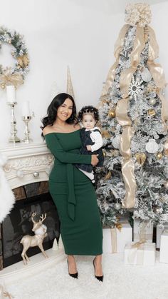 a woman in a green dress is holding a baby near a christmas tree and fireplace