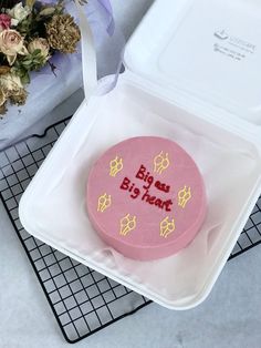 a pink cake with writing on it sitting in a plastic container next to a bouquet of flowers