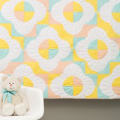 a teddy bear sitting on top of a white chair in front of a colorful quilt