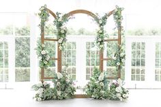 an arch decorated with flowers and greenery stands in front of two large windows that look out onto the outdoors