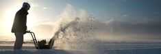 a man standing next to a snow blower on top of snow covered ground with the sun in the background