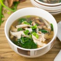 a white bowl filled with soup and greens on top of a wooden table next to a plate