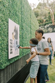 a man and young boy are painting a hedge on the side of a wall that says pin the tail on dad