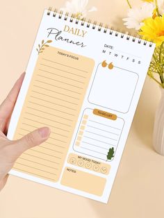 a hand holding a daily planner on top of a table next to flowers and a vase