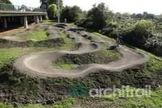 a man riding a bike down a dirt road next to a lush green hillside under a bridge