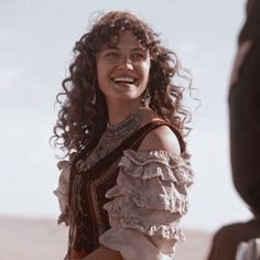 a woman with curly hair smiles while standing next to a man in a desert setting