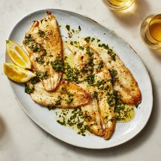 a white plate topped with fish covered in herbs and lemon wedges next to two glasses of wine