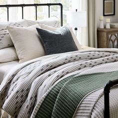 a bed with pillows and blankets on top of it next to a night stand in front of a window